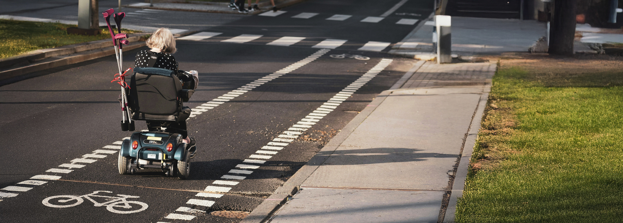 En person som använder elrullstol färdas på en cykelväg längs en gata. Personen har kryckor fästa på rullstolen och är på väg mot ett övergångsställe.